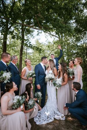 Wedding party gathers under trees for outdoor celebration featuring bridesmaids in blush and groomsmen in navy suits.