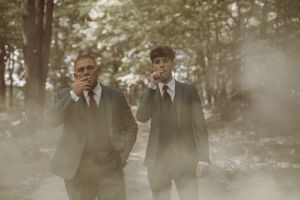 Two people in dark suits pose in a misty forest setting.