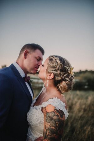 A romantic wedding portrait showing a bride with intricate braided hair and arm tattoo in a lace dress at sunset.