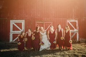 Bridal party in burgundy dresses poses together against rustic red barn doors at sunset.