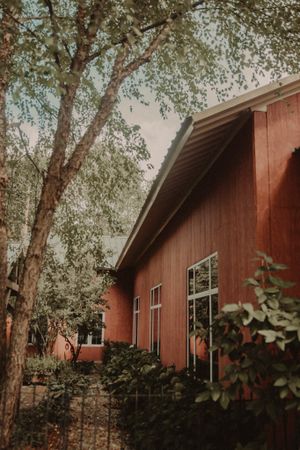 Red rustic building with white trim nestled among tall trees and lush landscaping.