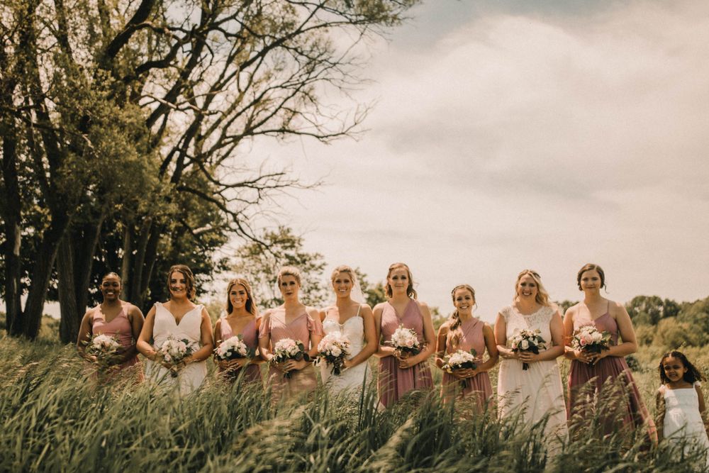 A wedding party poses in a line against a backdrop of tall trees and a cloudy sky in vintage-toned photography.