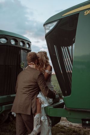 Romantic couple embraces in front of two large green tractors during a rustic farm wedding photoshoot.