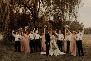 Wedding party celebrates with raised arms against a background of willow trees at sunset.