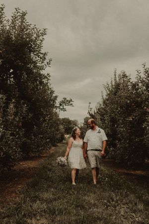A romantic couple walking hand in hand through an apple orchard on an overcast day.
