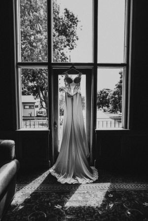 A white wedding dress hangs elegantly in a window frame, silhouetted against natural daylight.