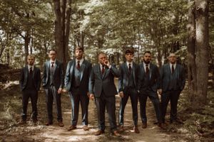 Group of groomsmen in dark suits walking through a wooded forest path.