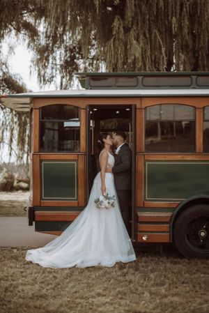 A wedding couple poses in front of a vintage trolley bus with willow trees draping overhead.