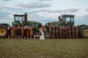 Wedding party poses between two large John Deere tractors in a rustic farm field setting.