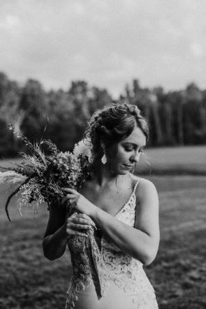 A black and white photo shows a bride in lace examining her wildflower bouquet in a meadow.