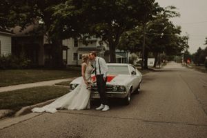 A wedding couple poses next to a classic vintage car on a residential street.