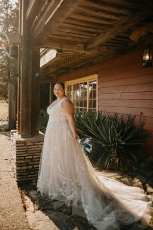 Bride in lace-detailed wedding gown with long train poses outside rustic venue.