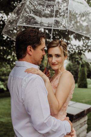 A couple shares an intimate moment under a clear umbrella in a garden setting.