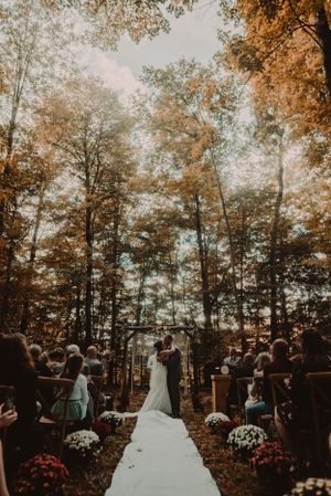 Outdoor forest wedding ceremony with white aisle runner and autumn foliage.