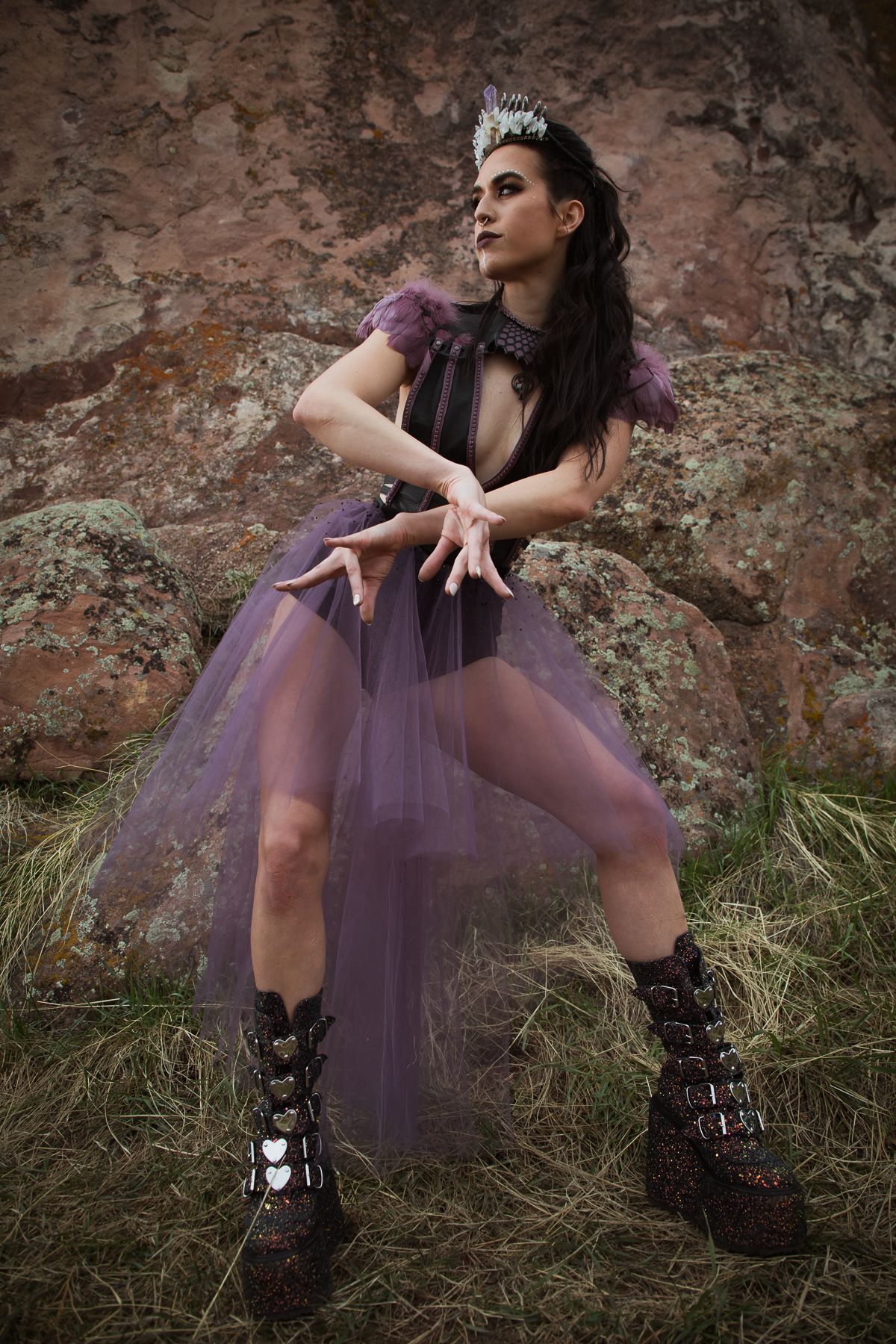 Alternative fashion model in purple tulle dress and combat boots poses against stone wall.