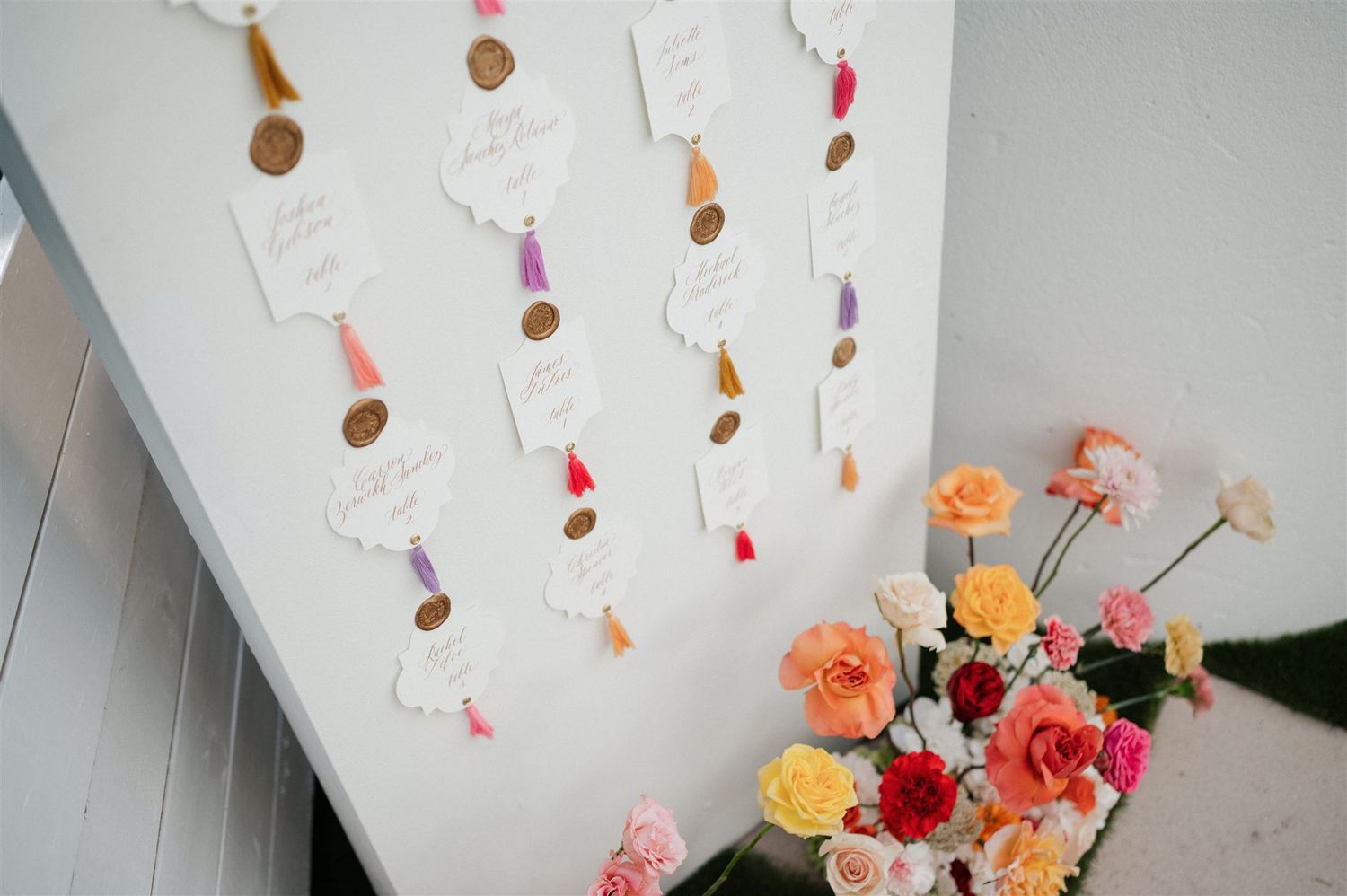 Colorful handmade seating chart with metallic spiral decorations hangs above a bouquet of mixed roses against a white wall.