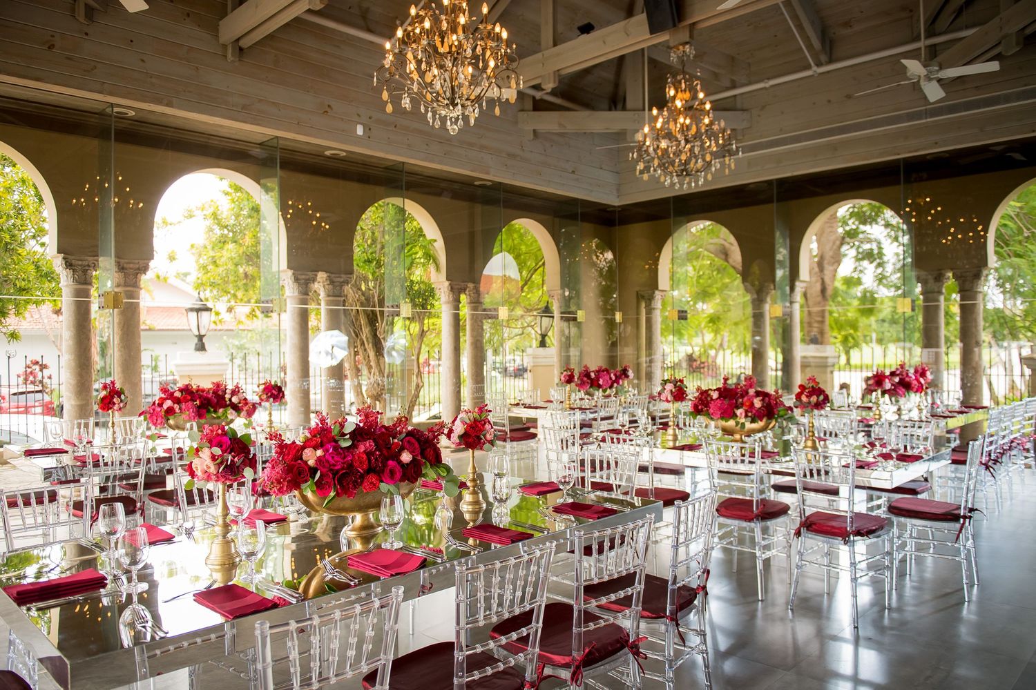 Elegant wedding reception venue with chandeliers, arched windows, and red floral table settings.