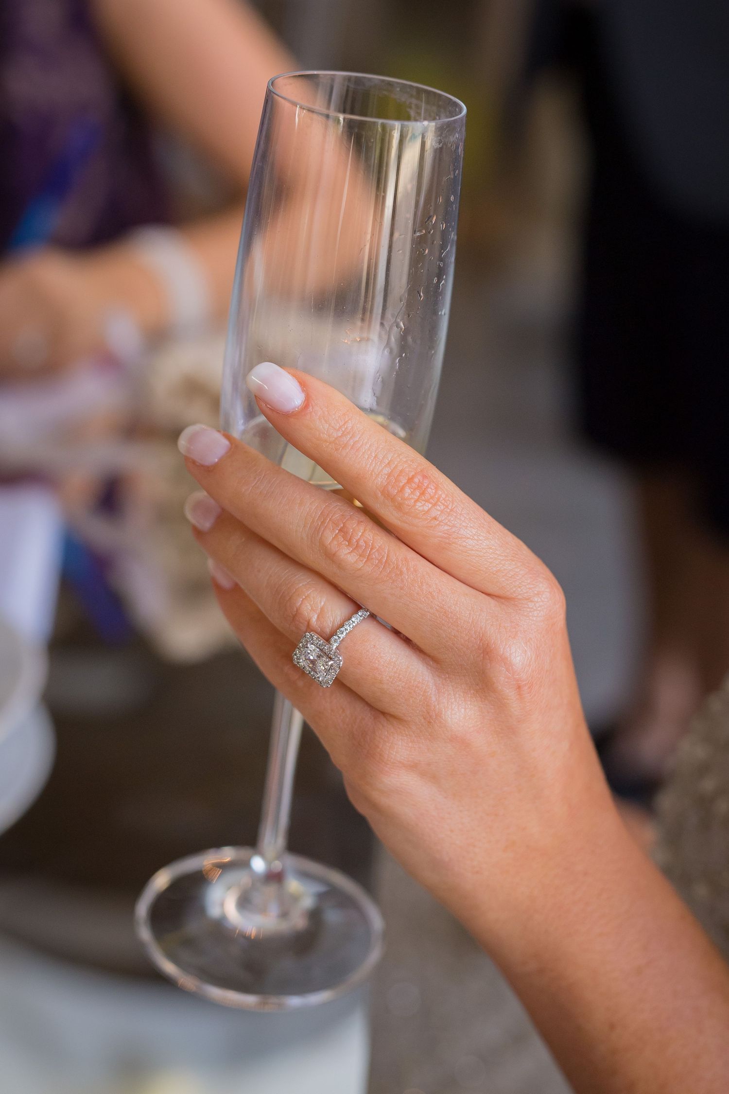 A manicured hand holding a champagne flute displays a sparkling diamond engagement ring.