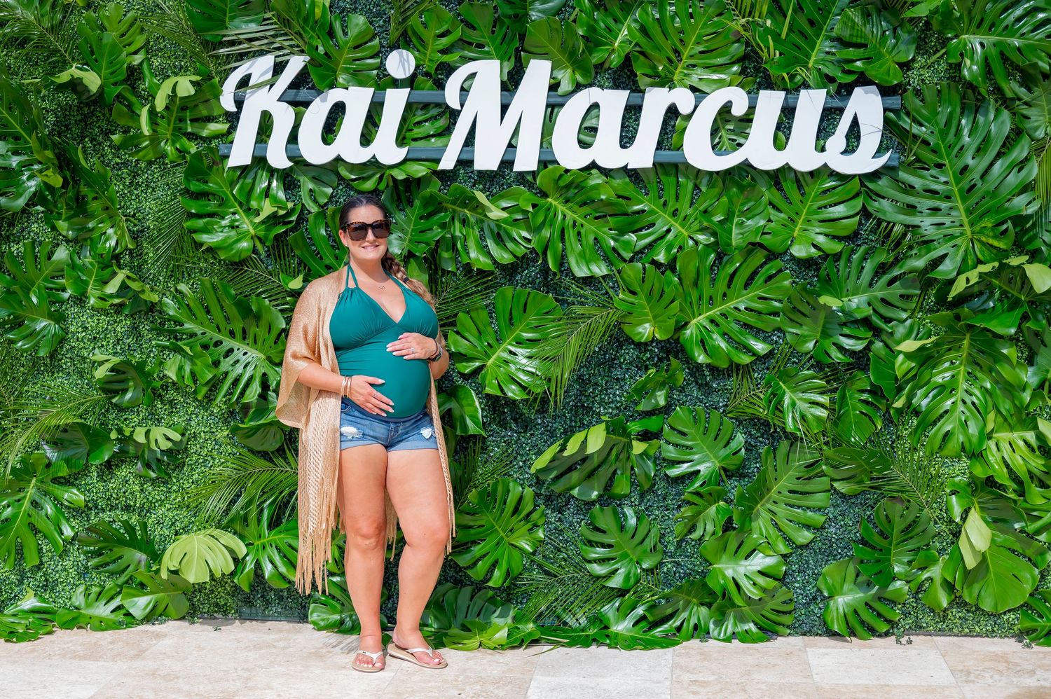 A mom-to-be poses in a teal swimsuit against a lush tropical leaf backdrop with a Kai Marcus sign above.