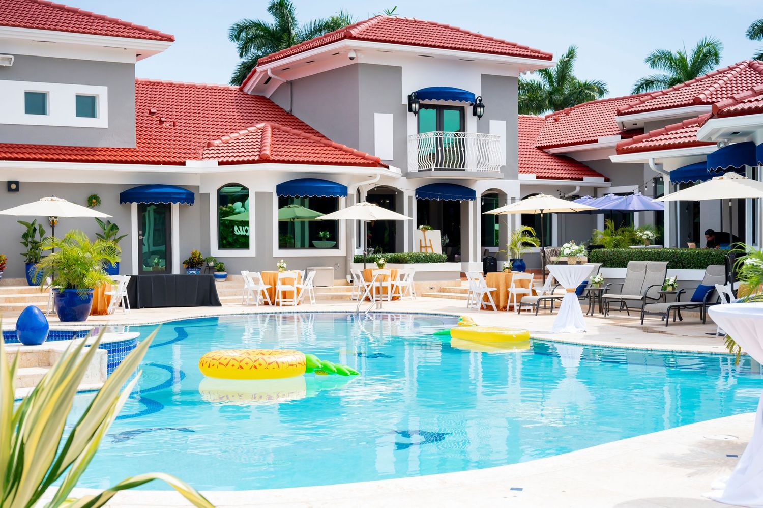 Luxury resort swimming pool with red-tiled buildings, blue umbrellas, and tropical palm trees surrounding the water.