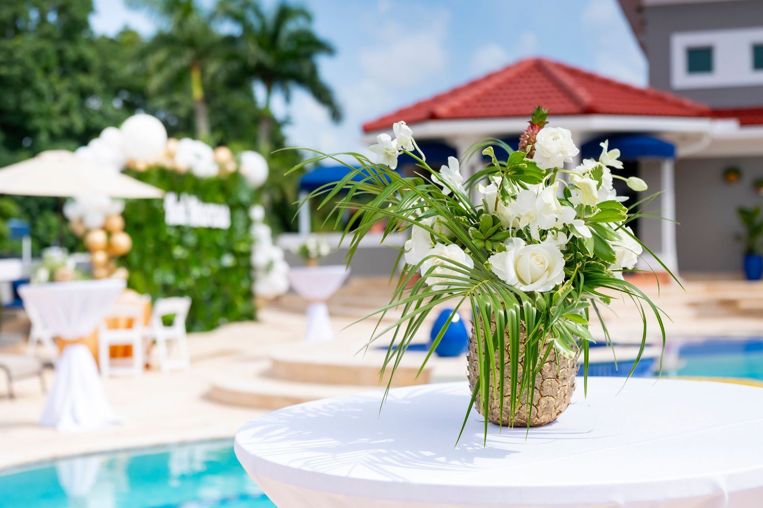 White floral centerpiece in pineapple vase on table by poolside with umbrellas and palm trees at tropical resort.