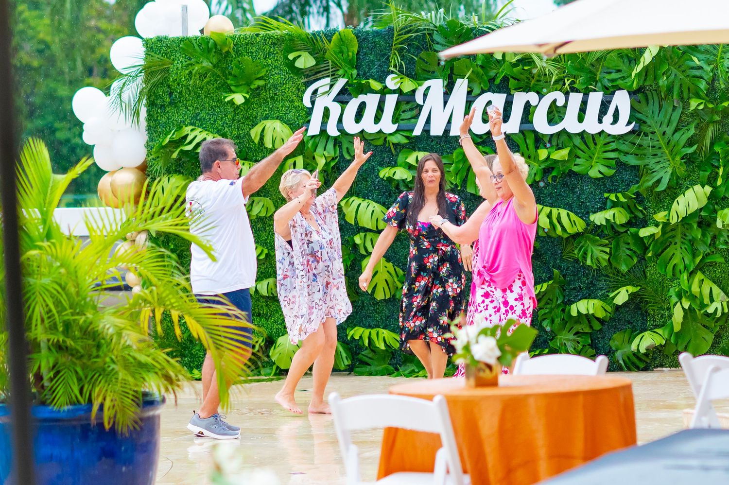 A group of people dance and celebrate together on a patio surrounded by lush green tropical plants.