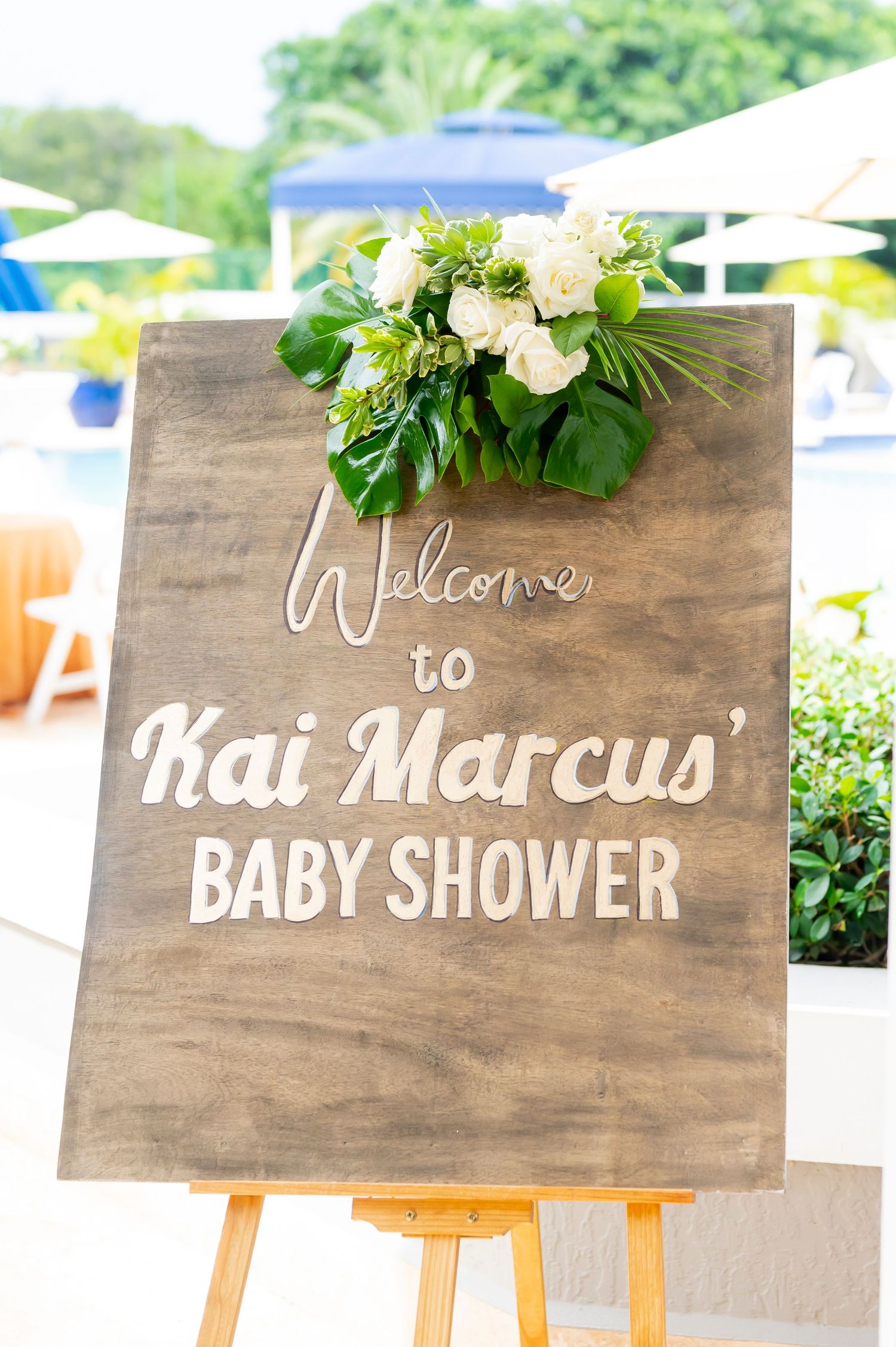 Wooden welcome sign for Kai Marcus' baby shower decorated with fresh white flowers and greenery on an easel stand.