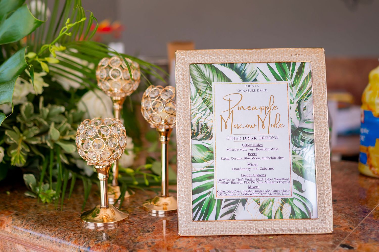 Drink menu with tropical palm leaves design sits in a gold frame next to decorative gold candlesticks.