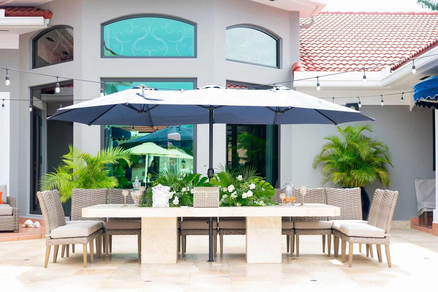 Outdoor patio dining area with beige chairs and tables under large navy umbrellas beside tropical palm plants.