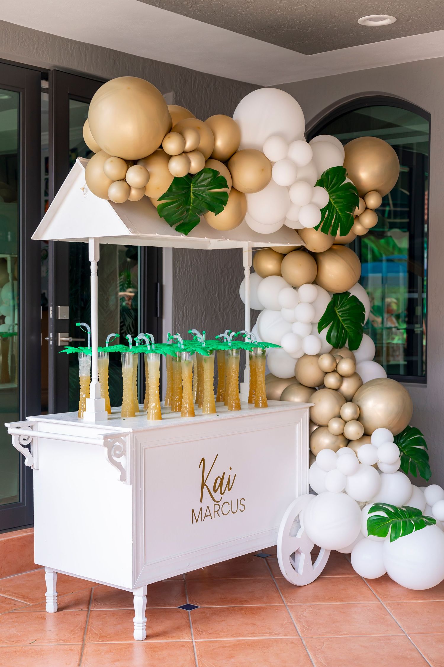 Elegant party bar cart with gold and white balloon arch garland and tropical monstera leaves against a textured wall.