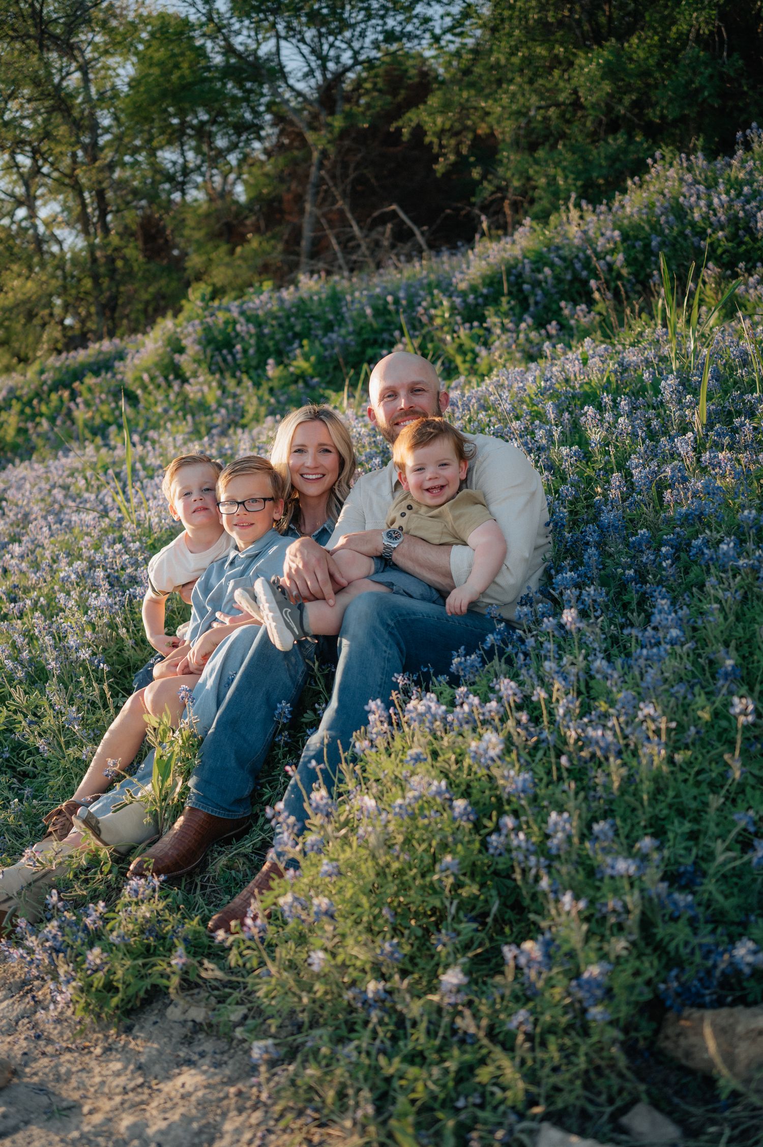 Family sits together on hillside covered in blooming blue wildflowers at sunset.