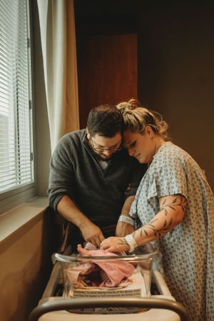 New parents lovingly attending to newborn baby in hospital bassinet by window.