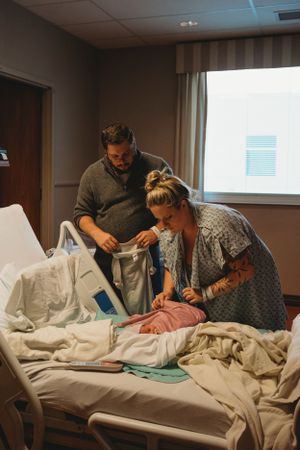 A couple in a hospital room attending to a patient in bed with medical equipment and curtains in the background.