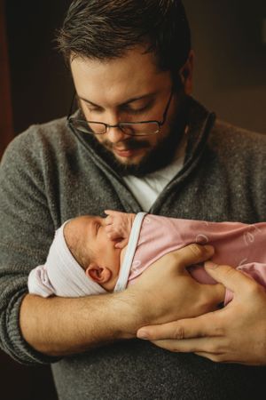 A person in a gray sweater tenderly holding a newborn baby wrapped in a pink blanket.