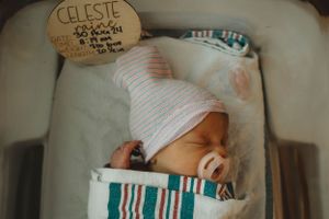 Sleeping newborn baby with pacifier in hospital bassinet with name tag visible.