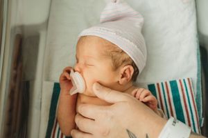 Newborn baby with white cap and pacifier resting in hospital bassinet.