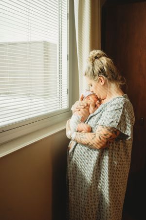 Person in hospital gown holding newborn baby while standing by window with blinds.
