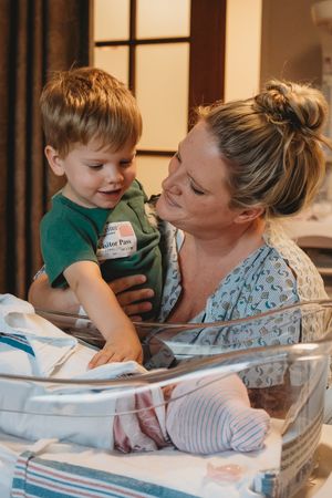 Mother and young child in green shirt bonding over newborn in hospital bassinet.