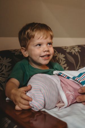 Child in green shirt holding a swaddled baby on a couch.