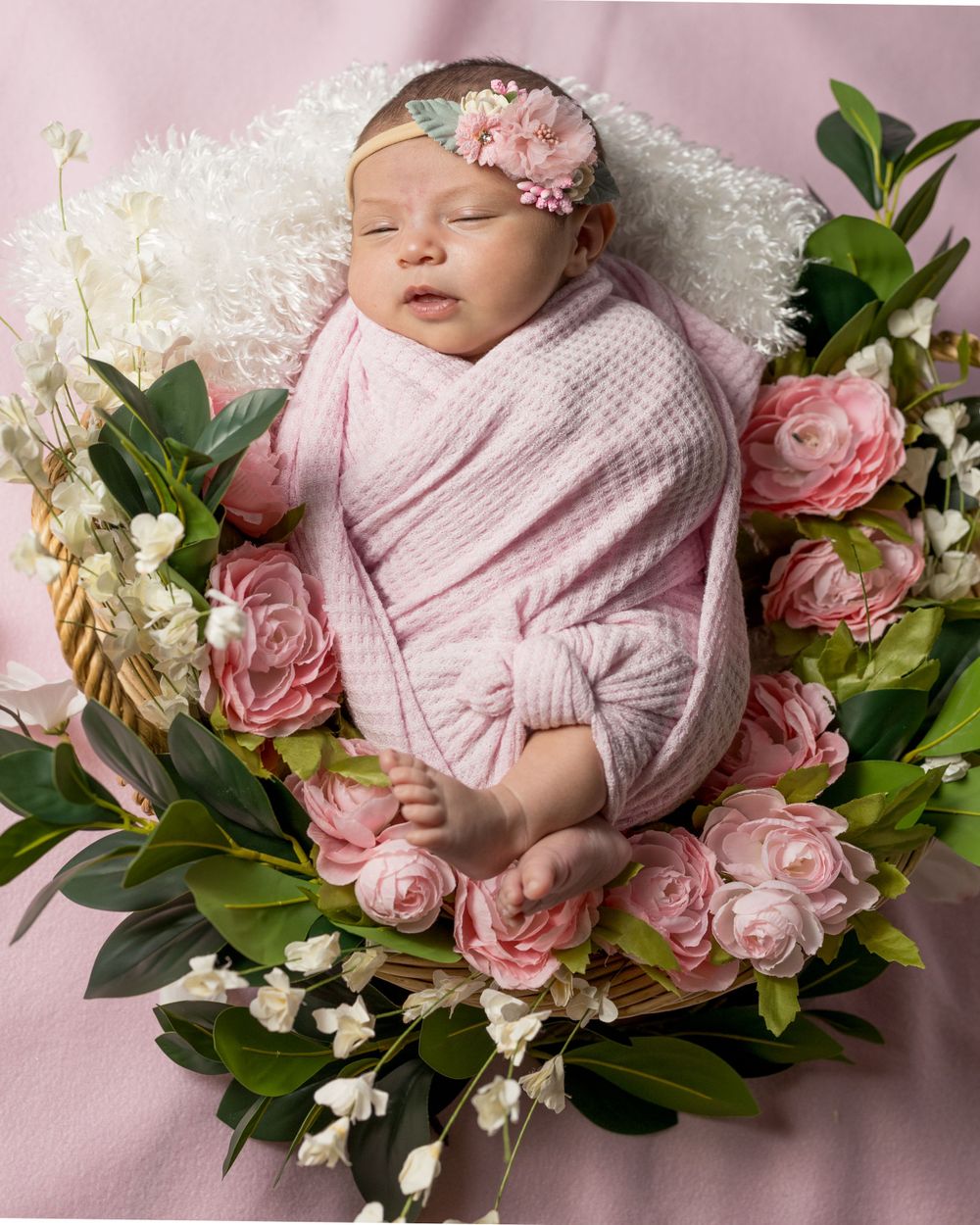 A sleeping newborn in pink wrap surrounded by pink roses and white flowers.