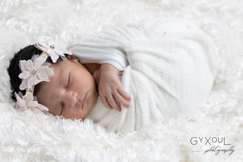 A sleeping newborn swaddled in white wearing a delicate floral headband on soft white fabric.
