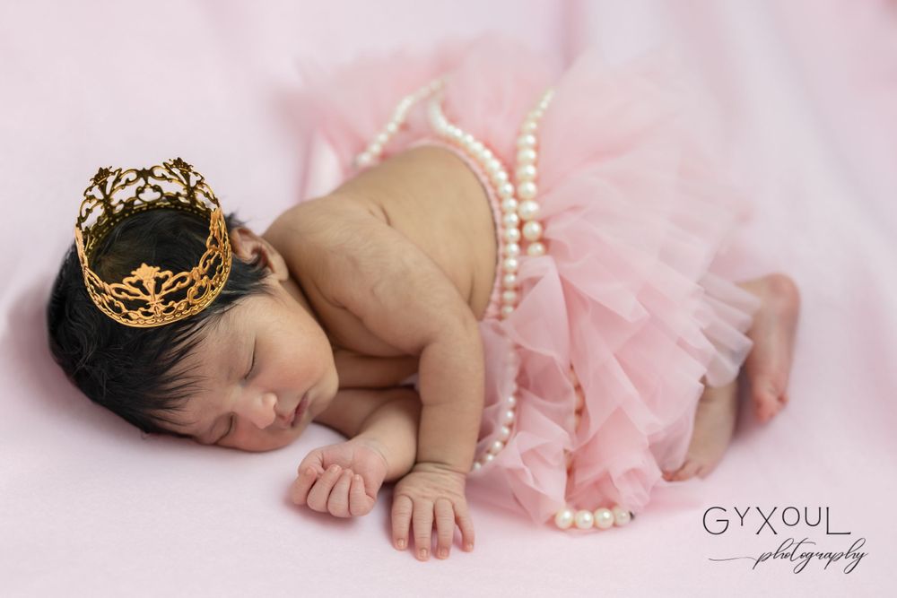 A sleeping newborn wearing a crown and pink tutu with pearls on pink background.