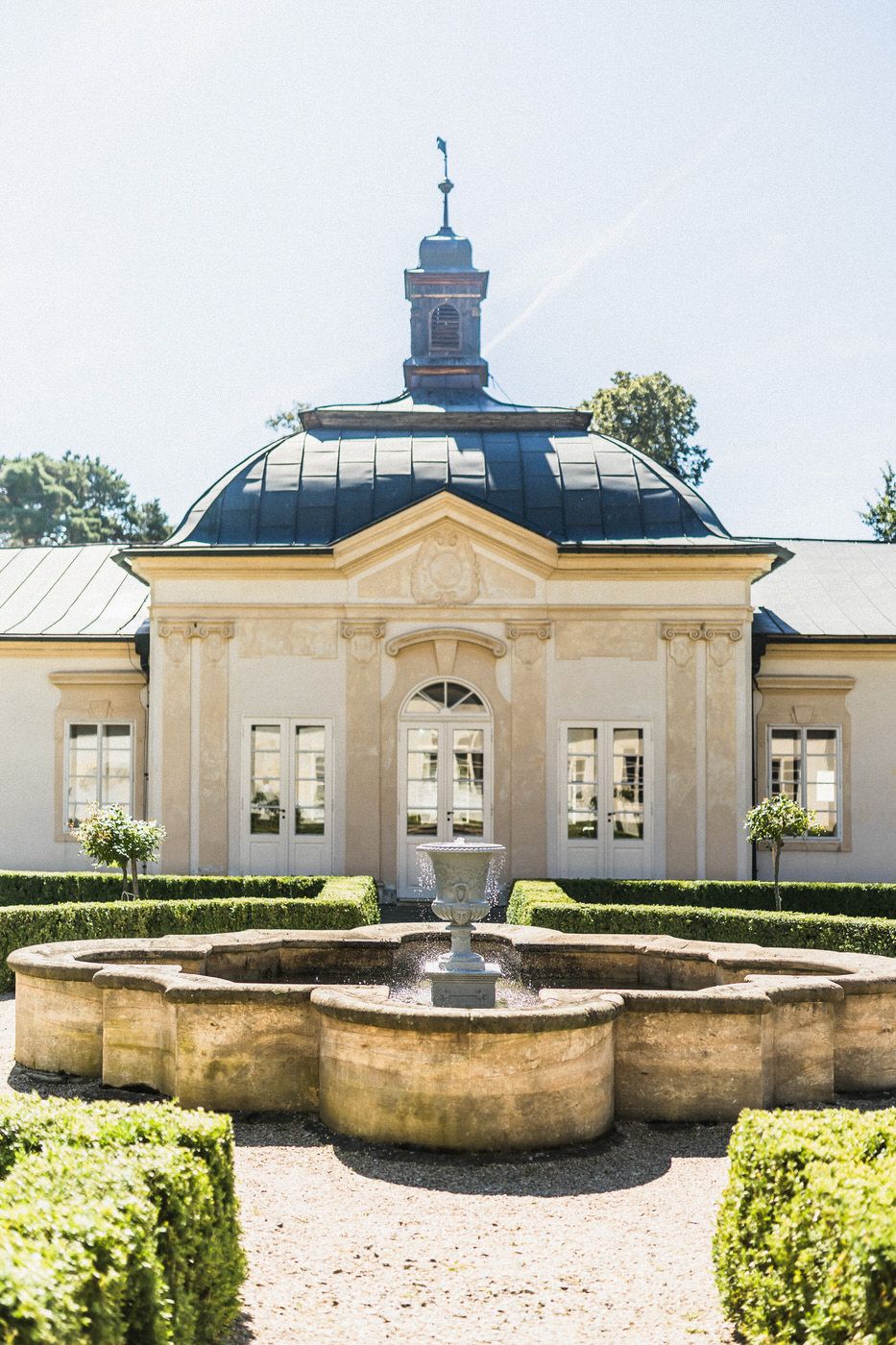 Elegant neoclassical building with domed roof and ornate fountain in manicured garden.
