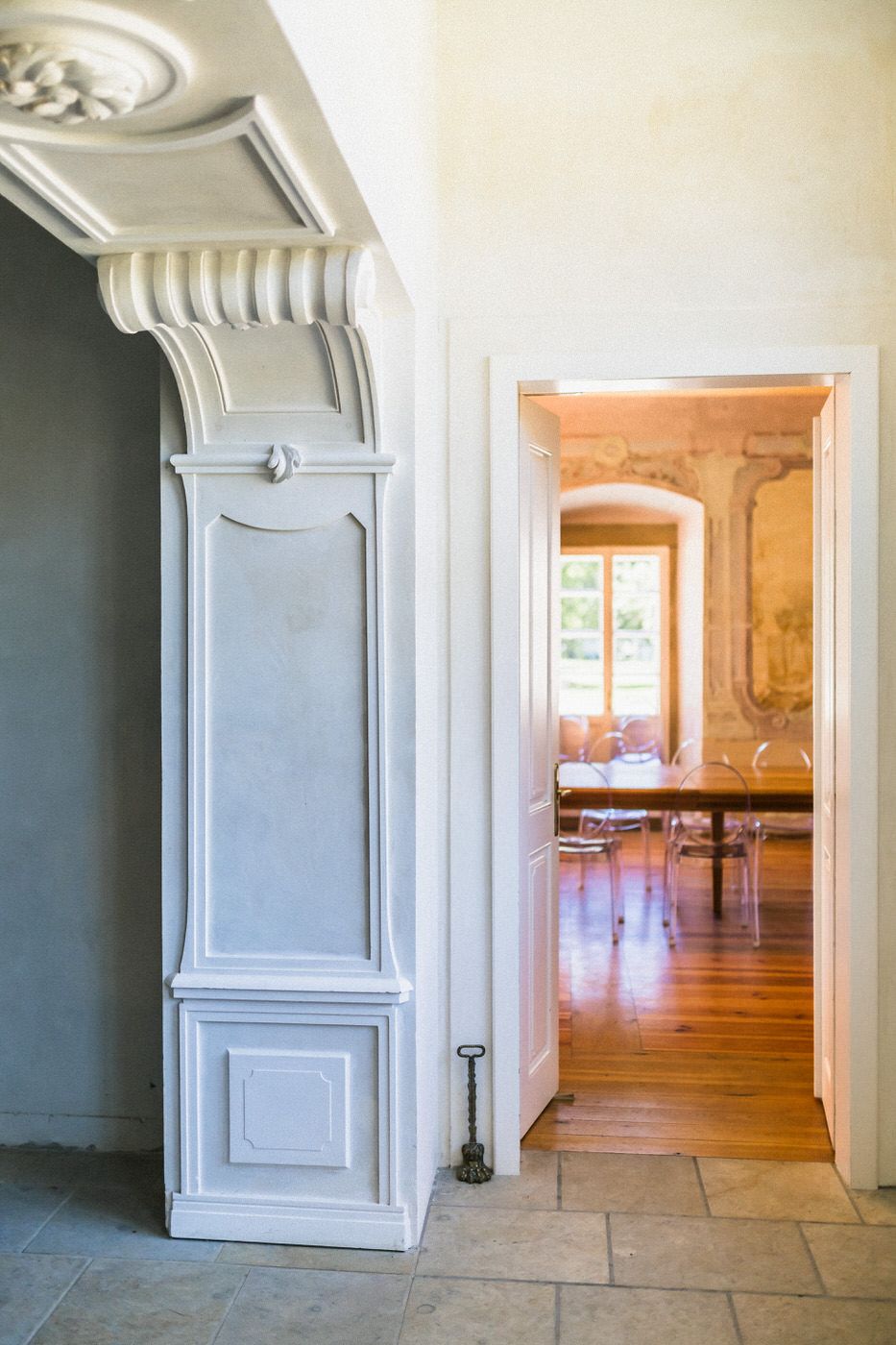 Interior doorway with ornate white molding leading to a sunlit room with hardwood floors.