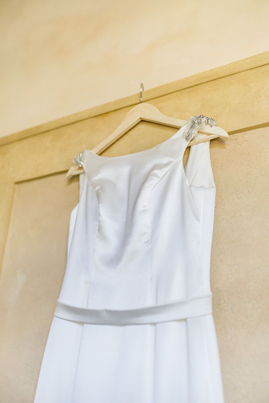 White wedding dress with embellished straps hanging on wooden hanger against beige wall.