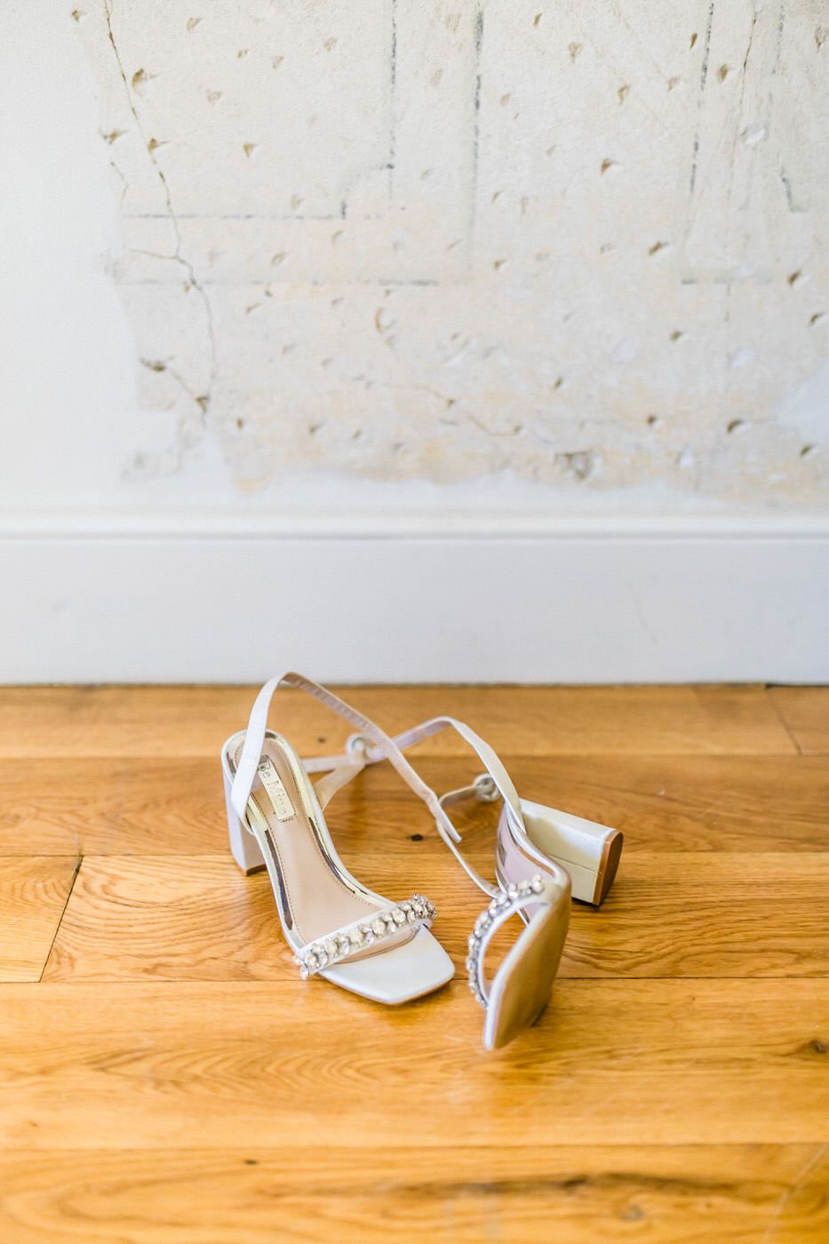 White wedding heels with crystal details on wooden floor against textured wall.