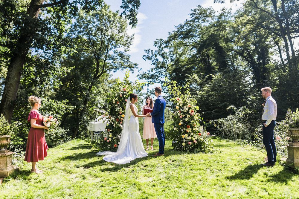 An outdoor wedding ceremony takes place under a floral arch in a sunlit garden setting.