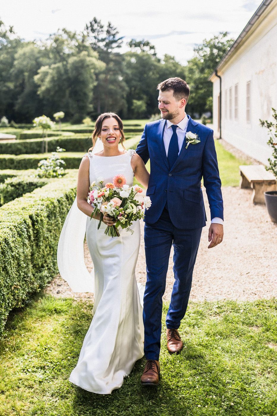 A couple strolls together through manicured gardens on their wedding day wearing classic formal attire.