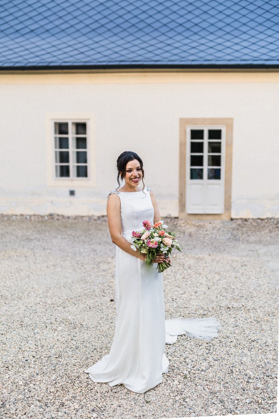 A bride in an elegant white sleeveless dress poses outdoors holding a beautiful bouquet against a stone building.