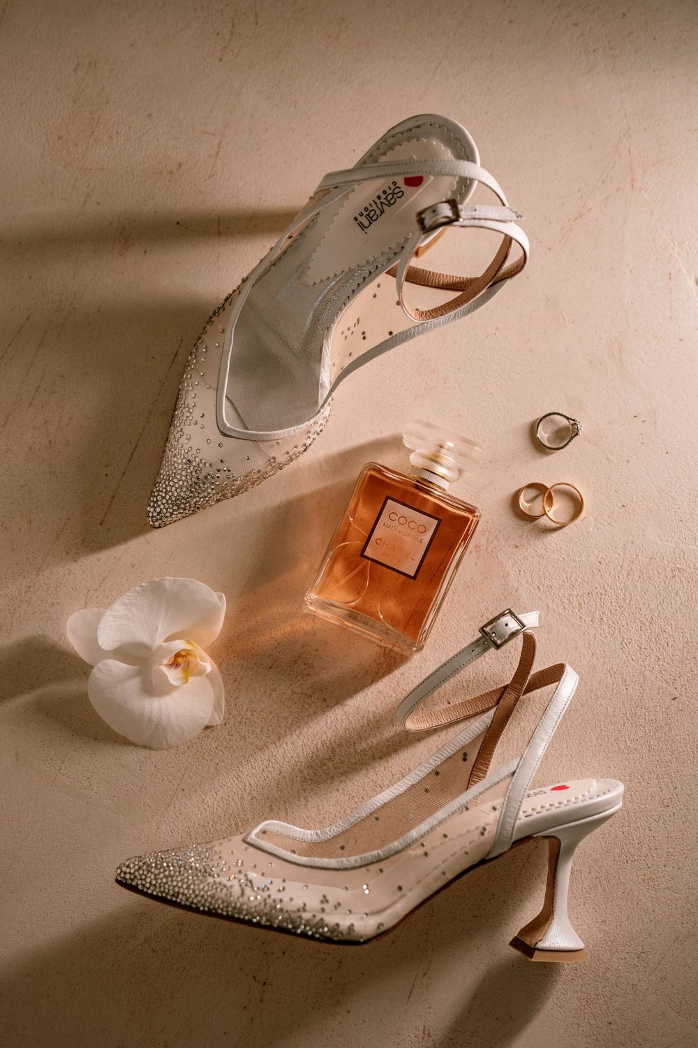 Elegant bridal shoes and perfume bottle arranged with wedding rings in soft beige lighting.